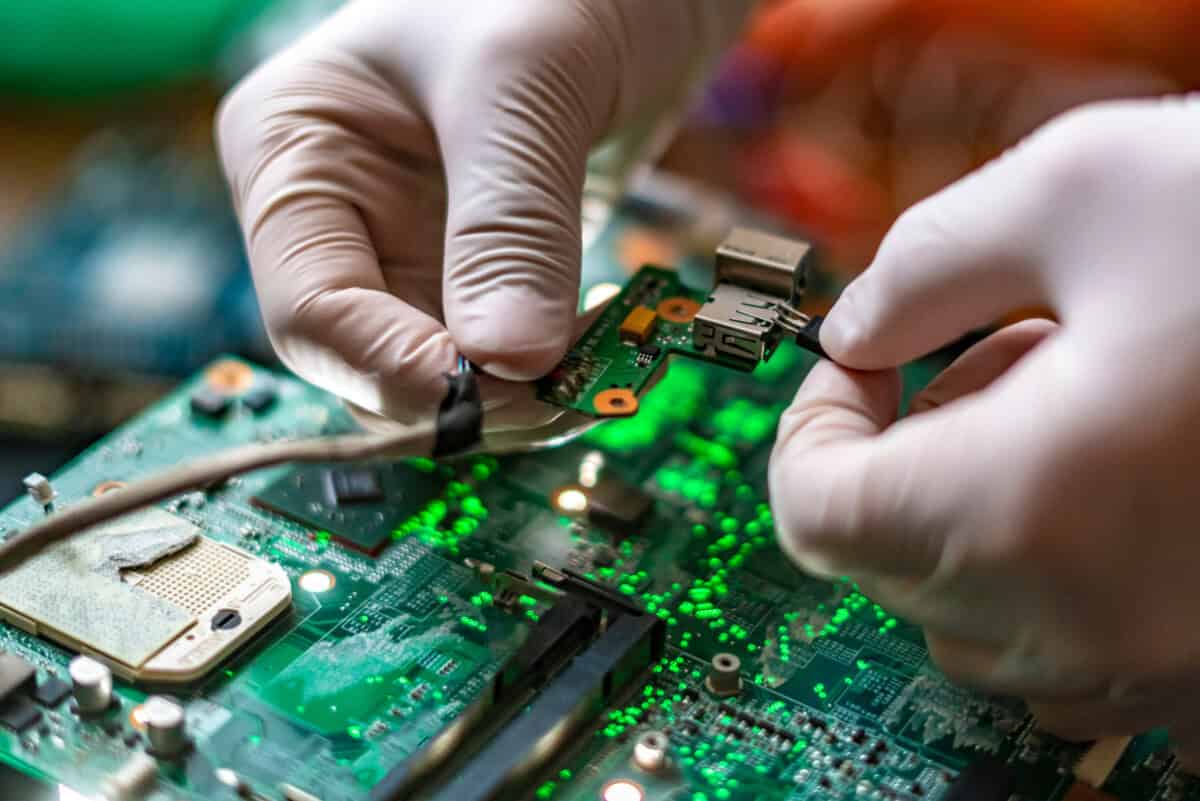Close-up of technician wearing white gloves performing a final assembly verification check on a green electronic circuit board (PCB) during quality control.