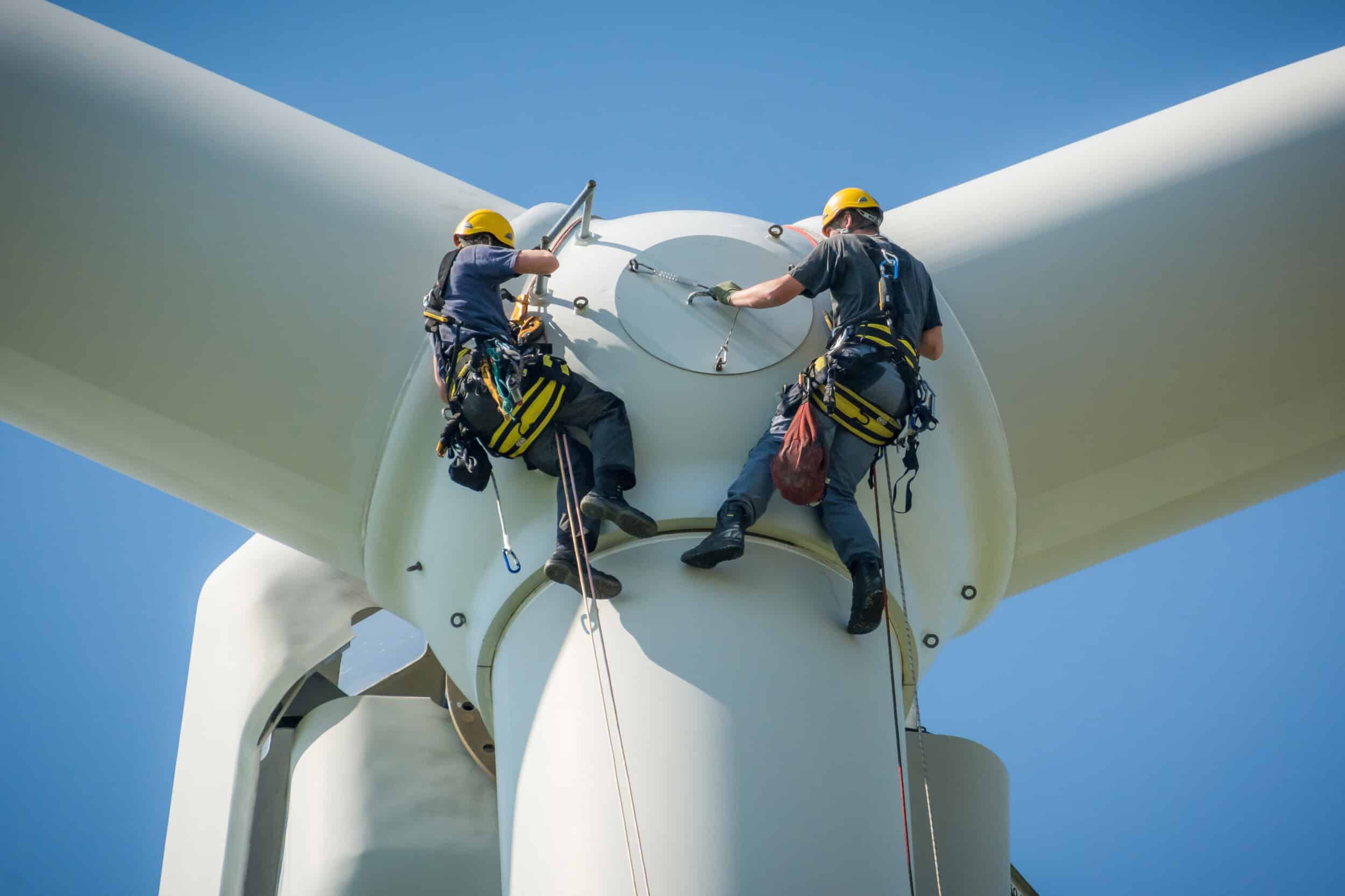 Two maintenance technicians in safety harnesses are suspended from a large wind turbine, performing an inspection or repair on one of its blades. This highlights the use of AI vision for enhancing worker safety, monitoring critical infrastructure, and detecting issues on wind farms in the energy and renewables sector.